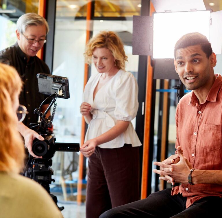 A man is explaining something enthusiastically to a woman sitting on a couch. A camera crew is set up behind the man.